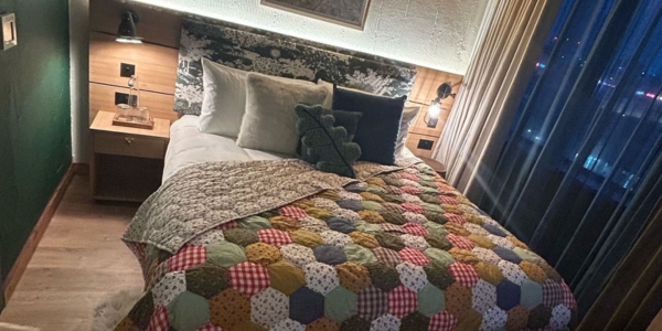 a double bed with a colourful patchwork quilt. The bed is a wooden floor in low lit room with night-time city views from a window next to the bed