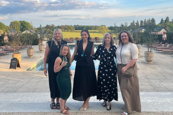five people pose for a photo in front of an outdoor pool that has ornate plant pots and sun loungers along two sides